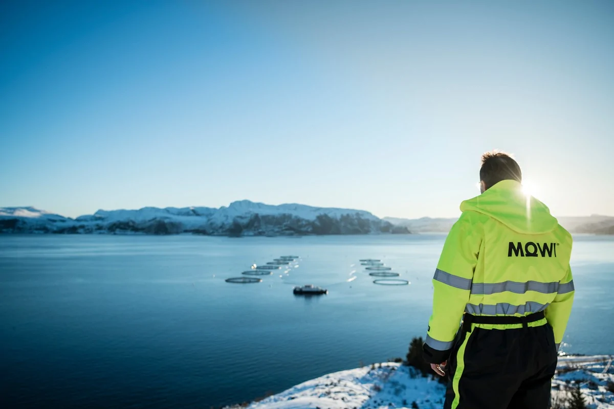 A man with neon green jacket with 'MOWI' written behind it, looking at the fishing boat on a blue sea