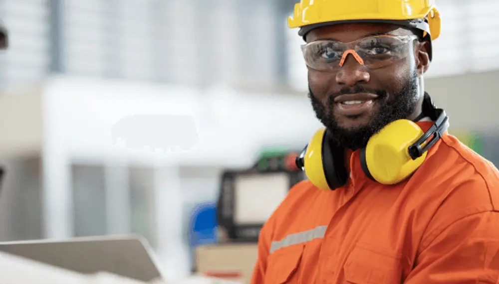 A construction worker wearing safety goggles and a pair of soundproof earphones around his neck smiles at the camera