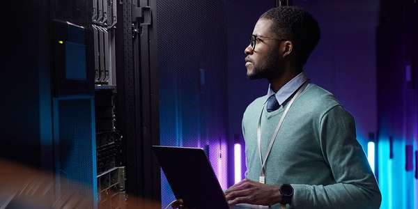 Ingeniero de datos afroamericano sosteniendo una computadora portátil mientras trabaja en la sala de servidores de supercomputadoras PartialIso Comms  