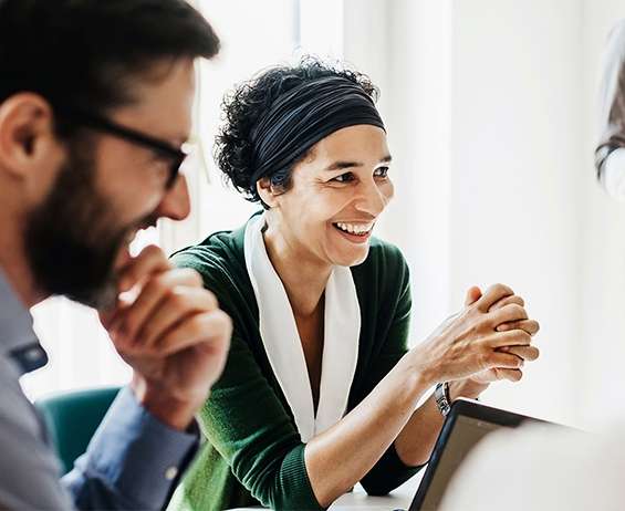 Two smiling colleagues – man in foreground, woman in focus – in an office at a meeting.