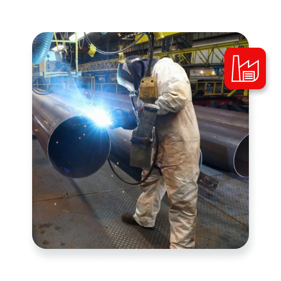 Worker in protective suit welding a large metal pipe in an industrial factory, with bright sparks and machinery in the background.