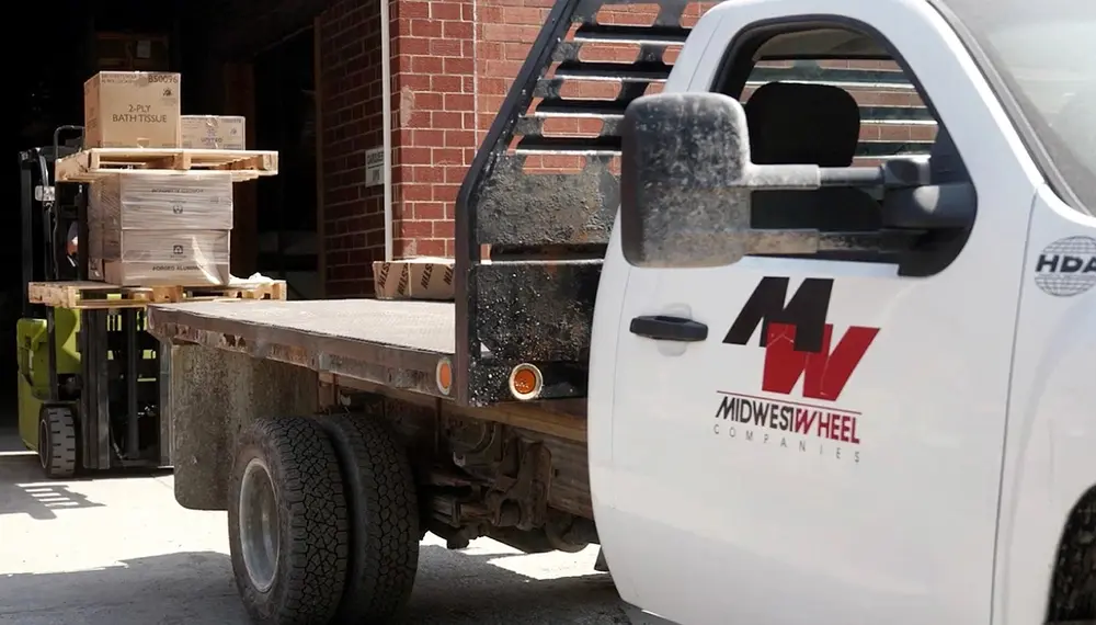 White truck with Midwest Wheel logo and forklift stacked with skids and boxes behind it