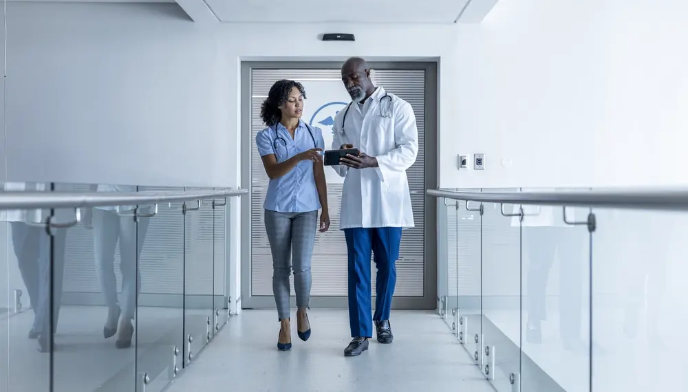 Female doctor walking in hospital corridor talking to a male coworker and looking at tablet