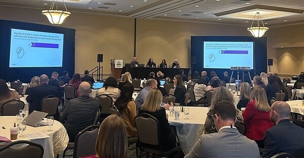 4 people seated at a table on a stage having a panel discussion in front of an audience at a conference. 