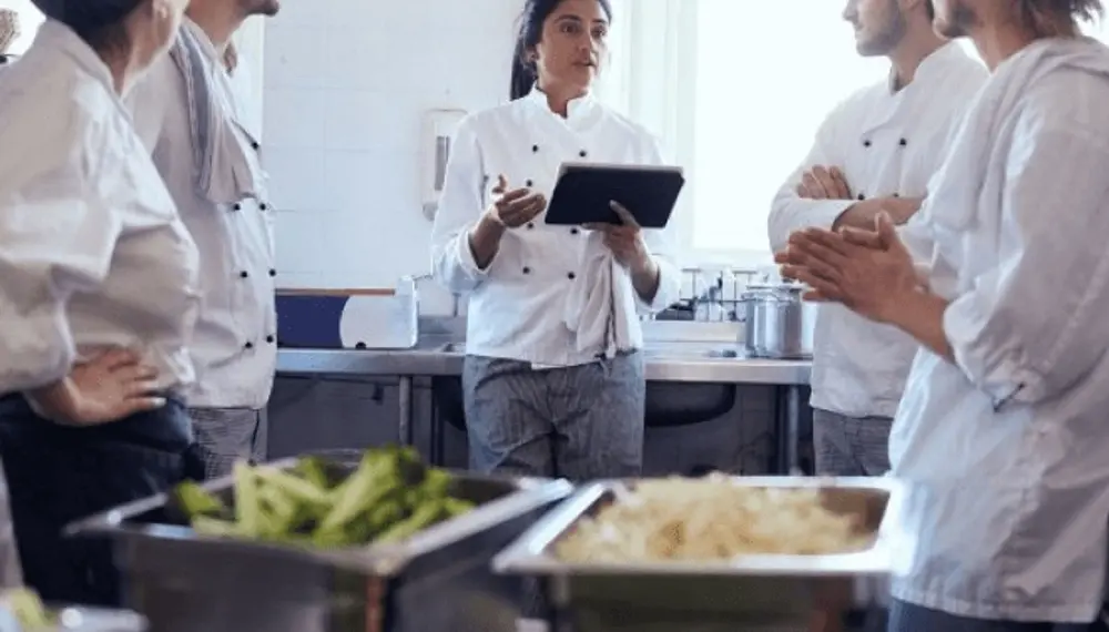 Multiple chefs in a kitchen listen to the head chef as she briefs them on the day's menu