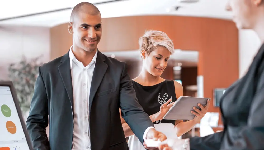 A guest hands over his card to a hotel front desk employee while his partner checks something on her tablet device