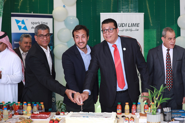 three men in suit jackets hold a knife preparing to cut a large white cake on a long table in celebration