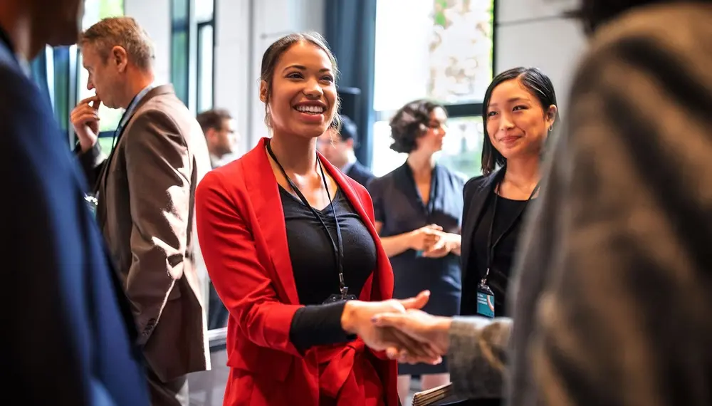 A business owner dressed in a red formal jacket shakes hands with her business partners at a conference