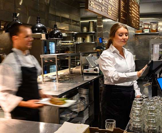 Prep cook walking by while server enters order data in a restaurant management system on a tablet.