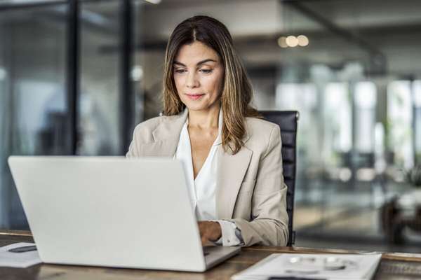 Woman in a light blazer and blouse sat at wooden desk focusing on her laptop