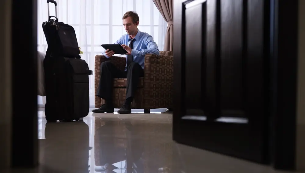Businessman in a hotel room with luggage sitting down on a chair on his tablet
