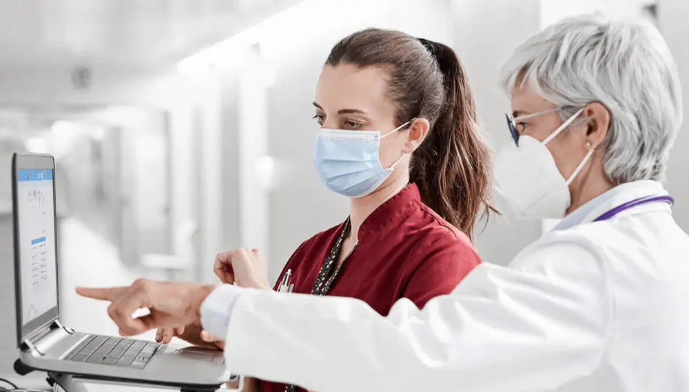 Two doctors wearing masks having a discussion in front of a laptop
